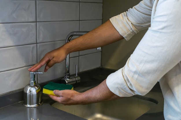 Hands of unrecognizable young Caucasian man with a scouring pad pouring soap to wash dishes