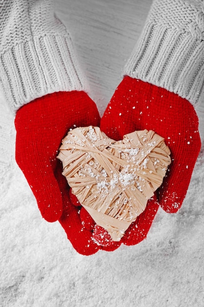 Hands in warm red gloves holding wicker heart on snowy background