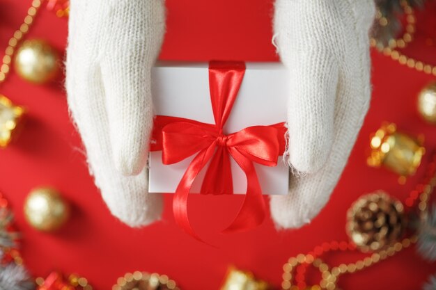 Hands in white knitted mittens holding a gift on a red background. White box with red ribbon. Sustainable holiday lifestyle. Christmas decorations