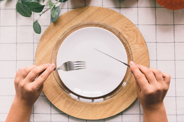 Hands with fork and knife, white plate on table.