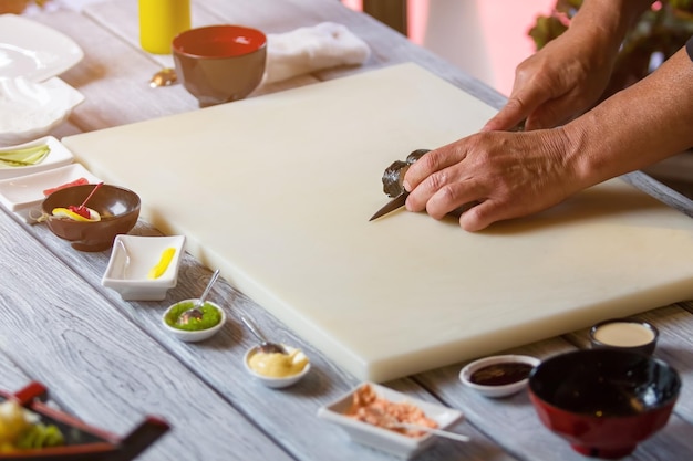 Hands with knife cutting sushi. Sushi on beige cooking board. Natural food from japanese cuisine. Tasty lunch cooked at cafe.