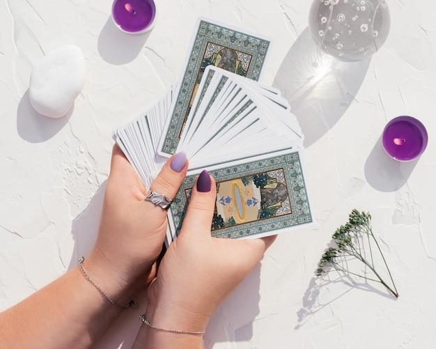 Hands with purple nails and rings hold deck of Tarot cards on white surface. Crystal ball, candles and stones, top view