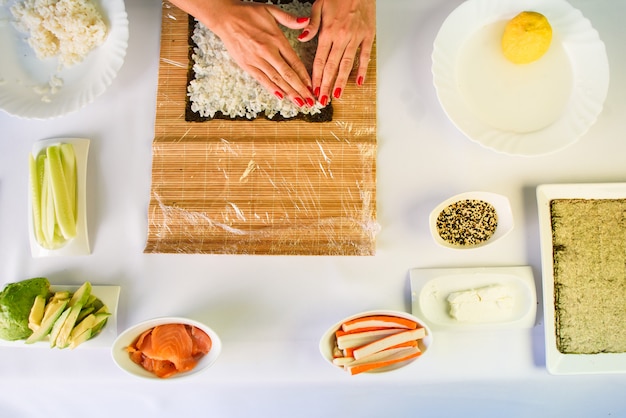 Le mani del cuoco unico della donna che riempiono i rotoli di sushi giapponesi con riso su uno strato dell'alga di nori.