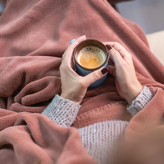 Hands of a woman holding a cup of hot coffee while warming up wrapped in a blanket for the winter