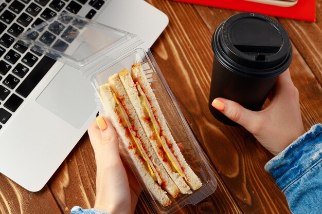 Hands of a woman holding sandwich above working table with laptop