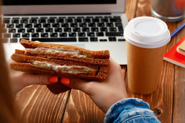 Hands of a woman holding sandwich above working table with open laptop