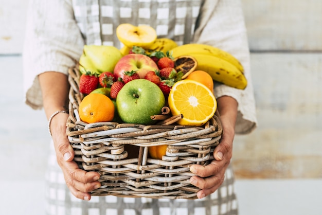 Hands of woman holding wicker basket full of colorful mixed fruit in abundance. Woman with variety of healthy fruit in basket. Fresh fruit collection for weight loss or healthy eating