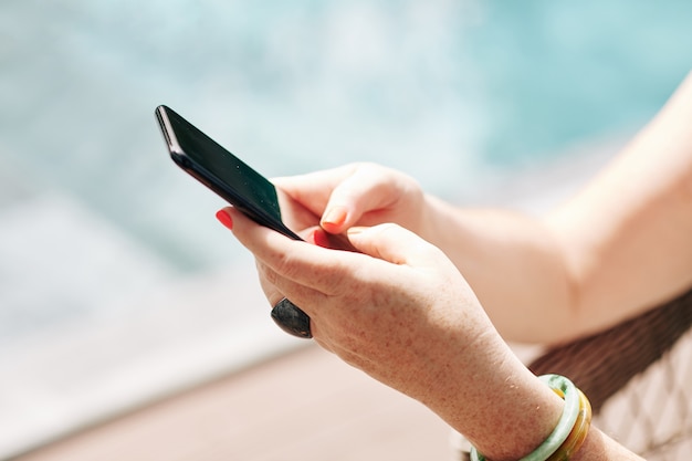 Hands of woman resting by swimming pool, texting and using mobile application