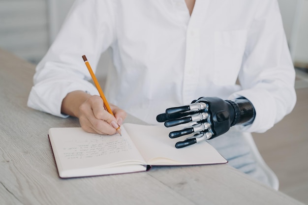 Hands of woman which is taking notes with pencil Cyber arm and healthy limb on the notebook