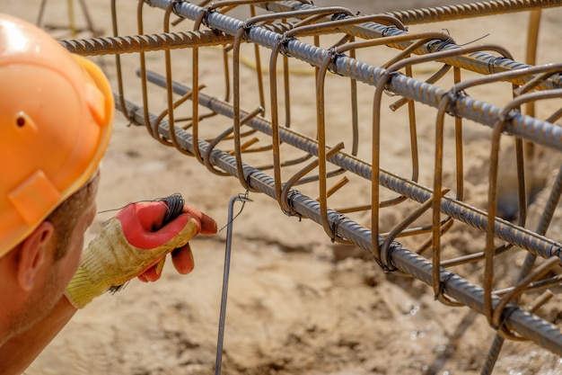 Hands of worker in protective gloves knit metal rods with wire for concrete reinforcement. Closeup view