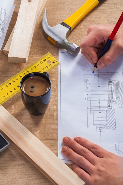 Hands of worker with pencil and blueprint on wooden table with coffee cup claw hammer ruler