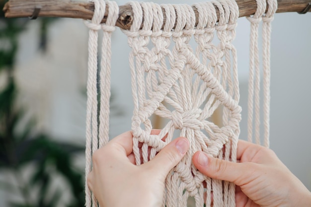 Hands working on a macrame piece suspended from a stick