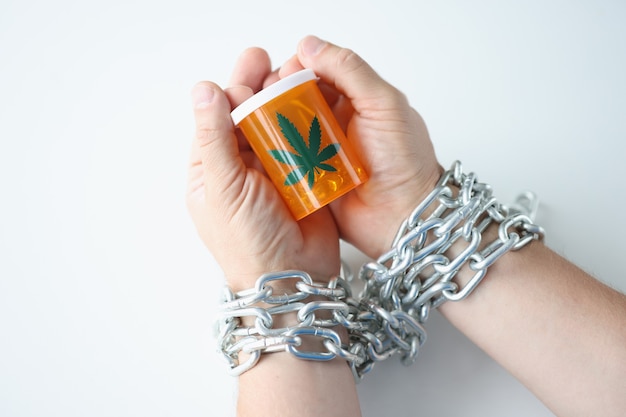 Le mani avvolte in una catena tengono un barattolo di dipendenza da marijuana e concetto di trattamento farmacologico