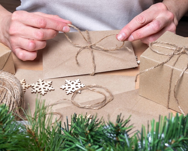 Foto mani che confezionano regali in uno stile ecologico, decorando una busta di carta kraft con una lettera di natale sul tavolo con regali di natale, spago e fiocchi di neve in legno. concetto di natale a rifiuti zero.