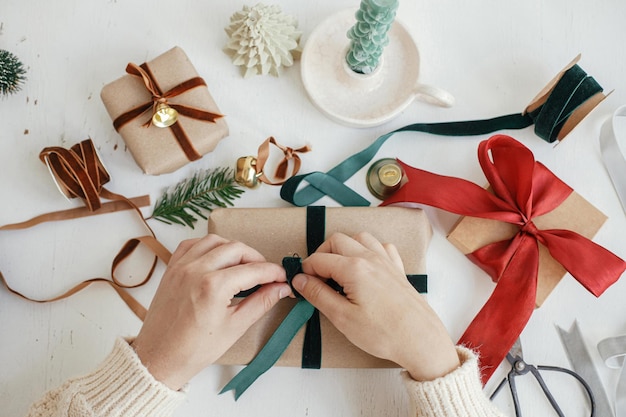 Photo hands wrapping stylish christmas gift flat lay person preparing christmas present with green ribbon and ornaments composition on white wood merry christmas aesthetic winter holidays