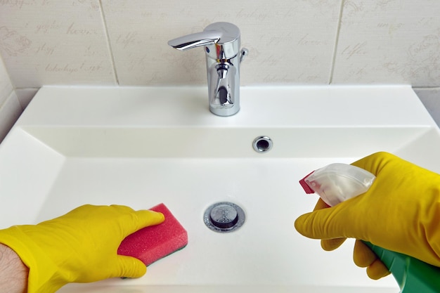 노란색 보호 장갑을 낀 손은 욕실의 위생 장비를 청소하기 위해 스폰지와 세제를 들고 있습니다.