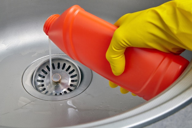 Hands in yellow rubber gloves pour sewer pipe cleaner down the kitchen sink drain