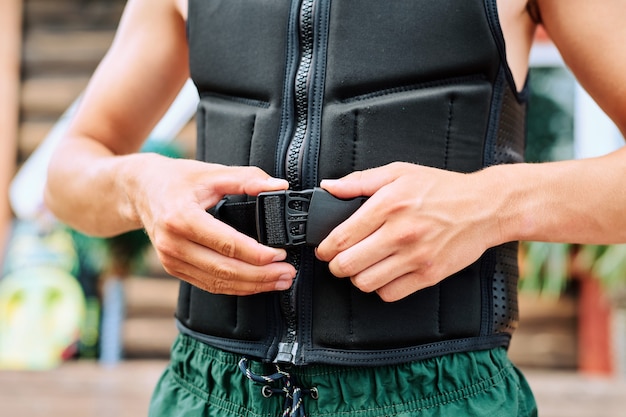 Hands of young active man fastening safety belt on black jacket against wooden house while going to have surfing training at summer resort