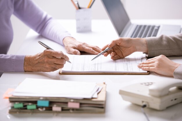 보험 문서를 가리키는 젊은 에이전트와 그녀의 서명을 넣을 고위 여성 고객의 손