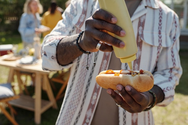 Hands of young black man putting mustard on top of hotdog with grilled sausage