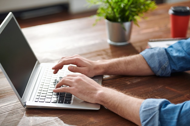 Hands of young businessman or designer touching buttons of laptop keypad while networking by table