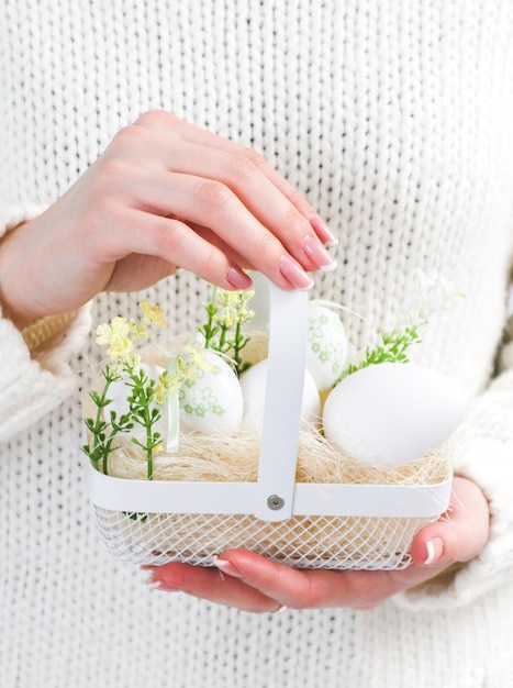 The hands of a young Caucasian girl hold a white metal basket with decorative Easter eggs