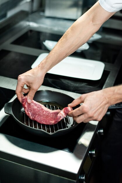 Mani di un giovane chef che mettono un pezzo di carne su una padella calda alla griglia con olio vegetale mentre sono in piedi vicino a una stufa elettrica e cucinano carne di manzo