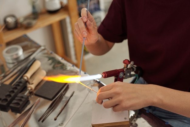 Mani di una giovane artigiana contemporanea seduta sul posto di lavoro e che brucia il pezzo con il fuoco mentre lo tiene sopra il bruciatore in officina