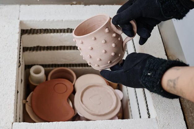 Hands of young female in black gloves taking clay cup created\
by herself