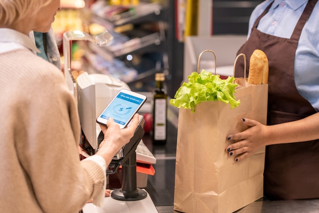 Hands of young female cashier or shop assistant holding paper sack with bread and lettuce leaves while customer paying for the goods