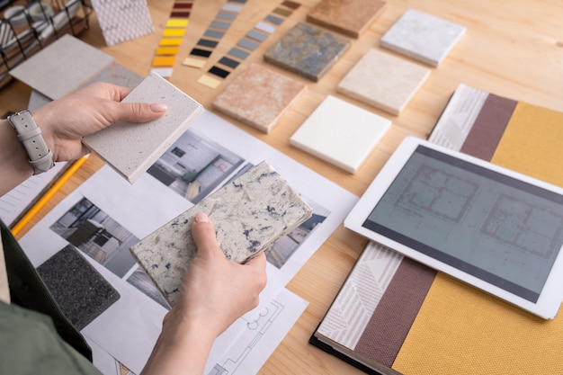 Mani di giovane designer femminile che tiene due campioni di piastrelle di marmo sul tavolo in legno con tavoletta digitale, foto di interni di casa ecc