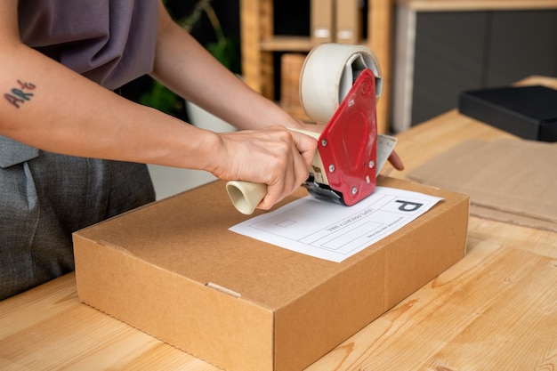 Hands of young female manager of online shop with cellotape gun dispenser sealing cardboard box with packed order of client by table