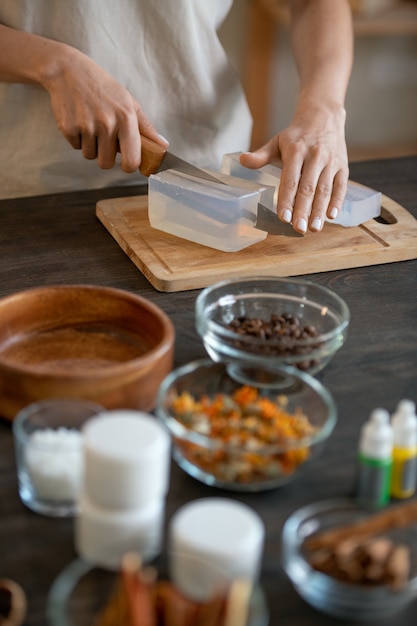 Mani di una giovane donna in piedi accanto al tavolo e che tagliano la massa di sapone duro su una tavola di legno mentre preparano prodotti cosmetici naturali a casa