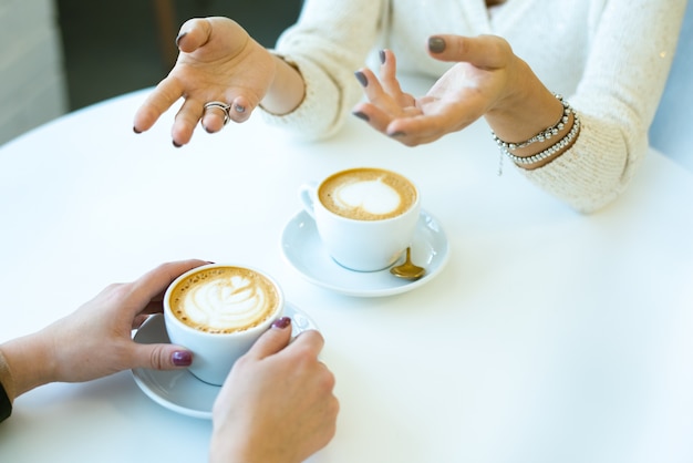 テーブルのそばに座ってカフェで新鮮なカプチーノを食べながら、目の前で友達と話している若い女性の手