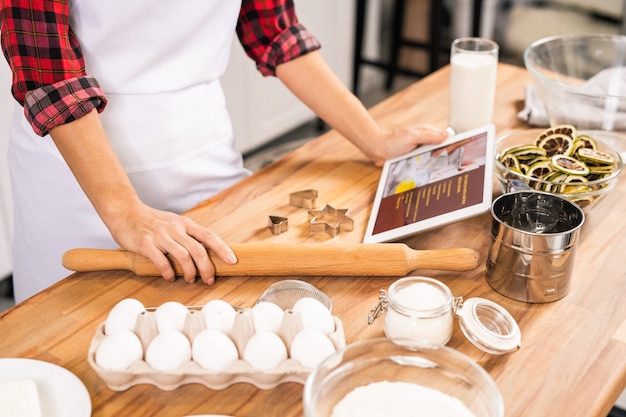 木製のテーブルのそばに立ってペストリーを調理しようとしている間、レシピで麺棒とタブレットを持っている若い主婦の手