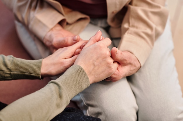 Hands of young kind woman holding those of mature female in casualwear while sitting next to her, supporting and comforting her in trouble