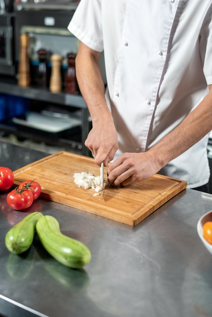 Hands of young male chef cutting fresh onions on wooden board surrounded by tomatoes and zucchini while cooking vegetarian lunch