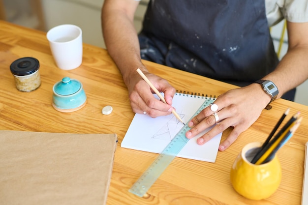 Hands of young man in apron sitting by table and making sketch with pencil