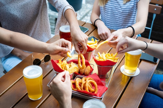 ジャンクフードとビールをテーブルの上で速く食べて揚げ物を食べる若者の手と不健康な食べ物の玉ねぎは素朴なチップとフライドポテトを鳴らします