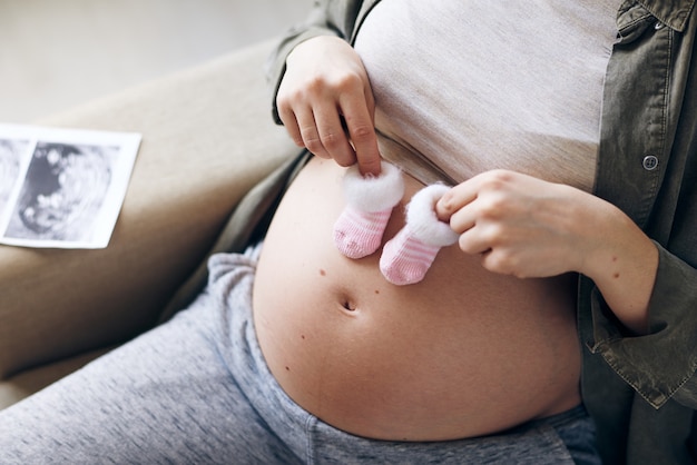 Hands of young pregnant female holding tiny socks for her future baby over belly while sitting in armchair or couch at home