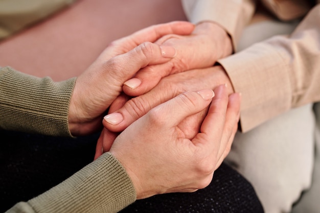 Hands of young supportive female counsellor holding those of mature woman while sitting next to her and comforting her in trouble