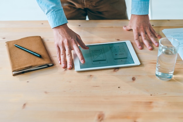 Hands of young website designer over program design on display of touchpad on wooden table during work