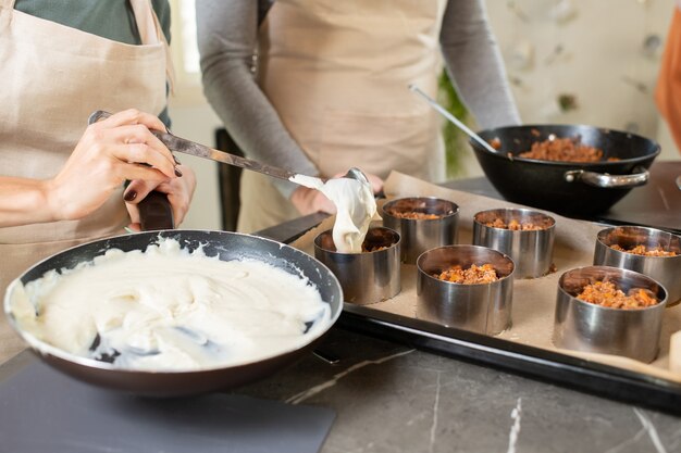 Hands of young woman in apron putting dough into metallic forms with fried minced meat before putting them into oven for baking
