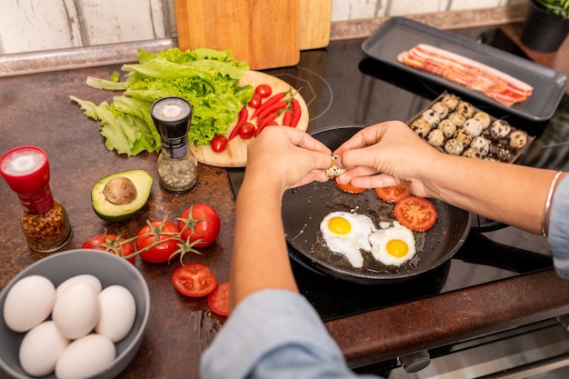 Mani di giovane donna che rompe le uova di quaglia fresche sulla padella calda mentre prepara la colazione con fornelli elettrici in cucina