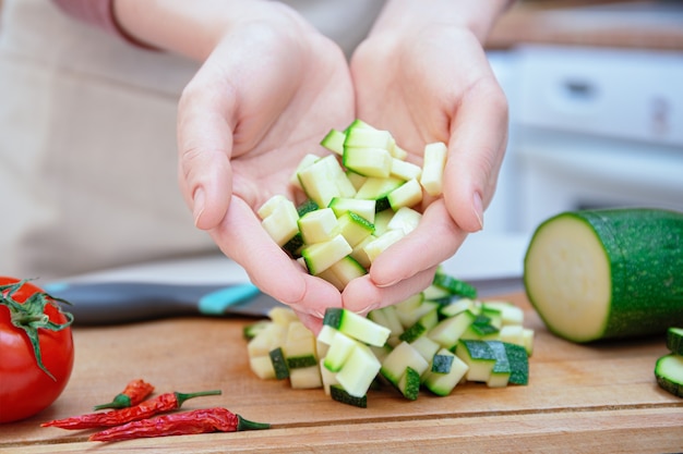 Hands of a young woman cut with a knife into cubes or slices of young zucchini cucumber on a wooden cutting board. Preparation of ingredients and vegetables before cooking. Healthy nutrition.