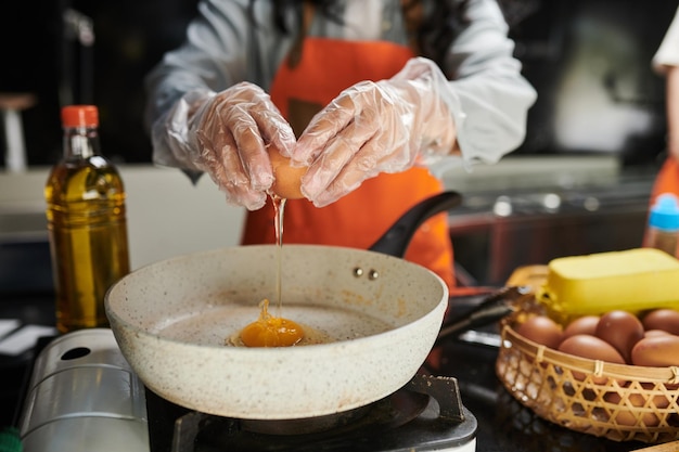 Hands of young woman in protective gloves breaking fresh egg on frying pan