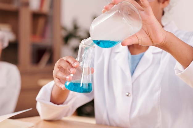 Hands of youthful mixed-race schoolgirl pouring blue fluid from bottle into tube with another chemical substance while experimenting