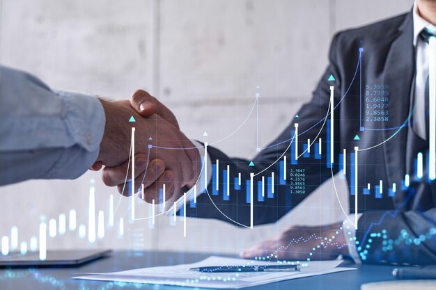 A handshake symbolize a capital market transaction to proceed profitable business in stock trading Financial hologram chart overt the table with the document