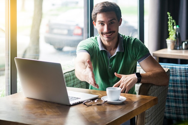 Handshake. Young happy businessman in green t-shirt sitting with laptop, toothy smile looking at camera and giving hand to greeting. business freelance concept. indoor shot near big window at daytime.