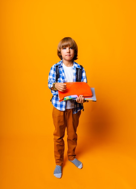 Handsome 7 year old boy with backpack holding books on yellow background for education concept